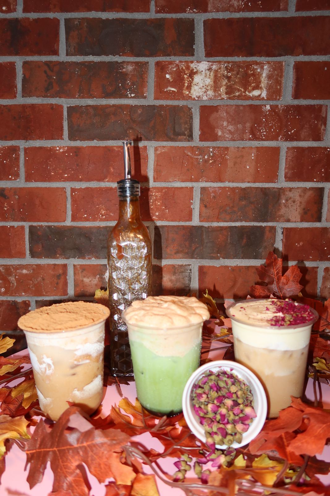 Three iced drinks on a pink table with autumn leaves, a bottle of syrup and a bowl of dried rosebuds, in front of a brick wall