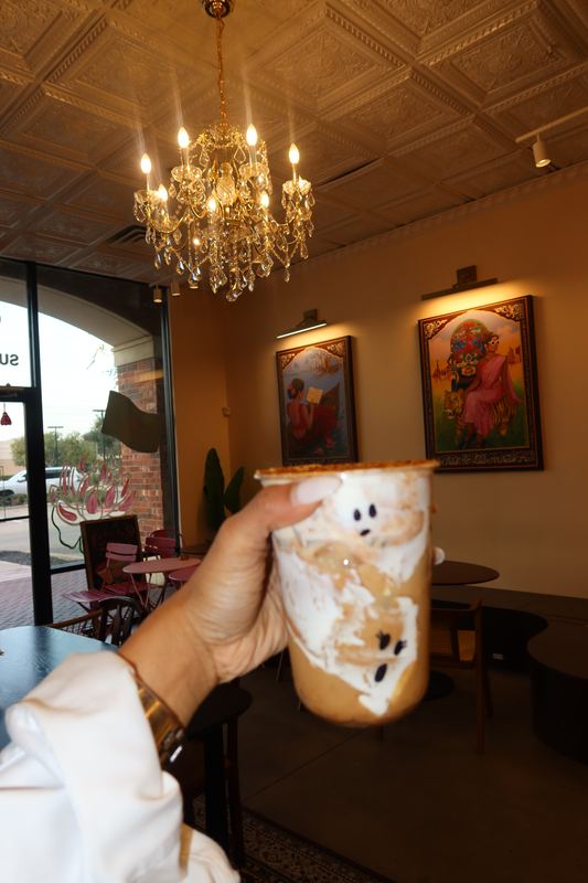 A hand holding a decorated iced latte inside Moharani, with a chandelier and framed art behind